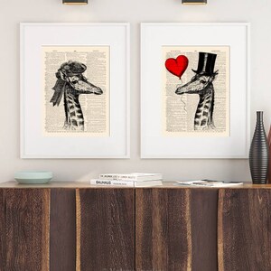 May include: Two framed prints of giraffes wearing hats. The giraffe on the left is wearing a hat with a ribbon. The giraffe on the right is wearing a top hat and holding a red heart balloon. The prints are on a white background with black text.