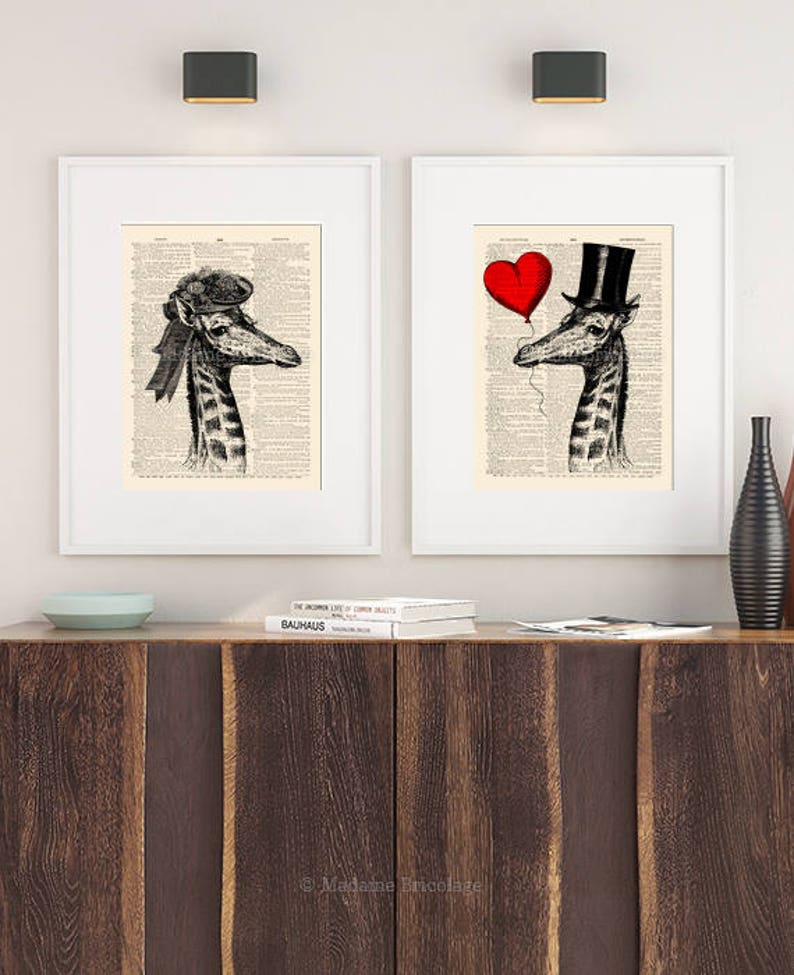May include: Two framed prints of giraffes wearing hats. The giraffe on the left is wearing a hat with a ribbon. The giraffe on the right is wearing a top hat and holding a red heart balloon. The prints are on a white background with black text.