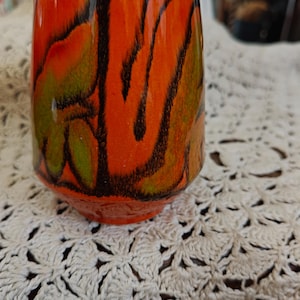 Może przedstawiać: Ceramiczny kubek z żywą pomarańczową podstawą, ozdobiony abstrakcyjnymi czarno-zielonymi wzorami. Kubek ma lekko rozszerzający się kształt i białą obwódkę. Projekt nadaje kubkowi niepowtarzalny, artystyczny wygląd.