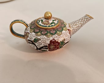 May include: A small, white teapot with a gold lid and intricate floral designs in shades of green, pink, and red. The teapot is made of cloisonné, a traditional Chinese enamelware technique.