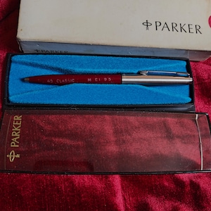 Può includere: Una penna Parker 45 Classic rossa e argento nella sua scatola originale. La penna è esposta in una custodia foderata in velluto blu, con il logo Parker visibile sulla scatola e sulla custodia. La penna ha il testo "45 CLASSIC" e "M £1 95".