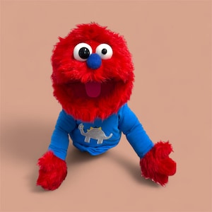 May include: A plush red puppet with large white eyes, a blue nose, and a pink tongue. The puppet wears a blue long-sleeved shirt with a silver dinosaur graphic. The puppet's hands and beard are red.