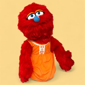 May include: A plush red puppet with large blue eyes, a blue nose, and orange dress. The puppet has a fluffy, textured appearance. The dress has white trim and a decorative pattern. The background is a solid yellow color.