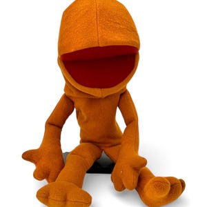 May include: A plush orange puppet with a wide open mouth and a red interior. The puppet has a hooded head, arms, and legs, and is sitting down. The puppet's features are simple and cartoonish.
