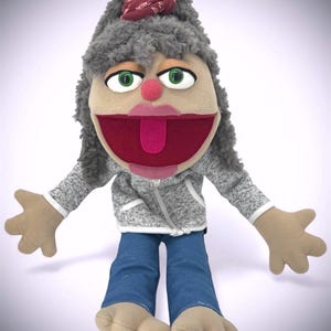 May include: A plush puppet with a light skin tone, gray hair, and green eyes. It wears a gray zip-up hoodie, blue jeans, and has a red bow on its head. The puppet's mouth is open, revealing a red interior and a pink tongue.