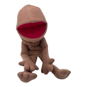 May include: A brown plush puppet with a large, rounded head and a wide, open mouth featuring a deep red interior. The puppet has long, slender arms and legs with three-fingered hands and feet, all crafted from a soft, textured brown fabric.