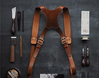 May include: A brown leather camera strap with adjustable buckles and a padded chest piece. The strap is laid out flat on a dark gray surface with various tools and supplies.