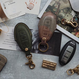 May include: Two leather key fob covers in olive green and brown, with a black Nissan key fob. The key fob covers have brass fittings and are stitched with contrasting thread. Crafting tools and materials are scattered around the key fobs.