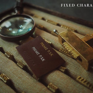 May include: A close-up of a leather label with the text "PIXAKE P.XK" embossed in gold. The label is lying on a surface with a magnifying glass and a brass tool with a row of metal characters.