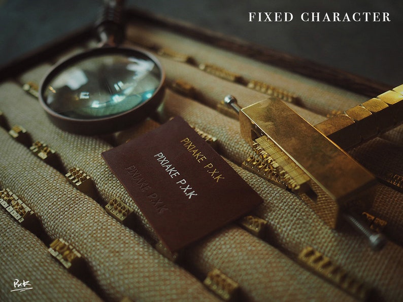 May include: A close-up of a leather label with the text "PIXAKE P.XK" embossed in gold. The label is lying on a surface with a magnifying glass and a brass tool with a row of metal characters.