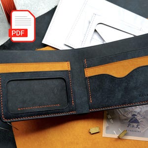 May include: A bifold wallet crafted from black leather with contrasting orange stitching and interior accents. The wallet features a clear ID window slot, multiple card pockets, and a bill compartment. It is displayed alongside leather scraps, a stitching chisel, needles, and paper templates.