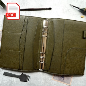 May include: An olive green leather binder featuring a six-ring silver metal mechanism, multiple interior pockets, and a side strap closure. The binder is surrounded by various metal and wooden crafting tools, including pliers and a chisel.