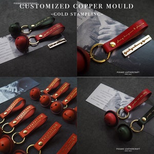 May include: A set of red leather keychains with gold-stamped lettering and a small bell charm. The keychains are arranged on a black surface. The text on the keychains reads "TATATATTOO".