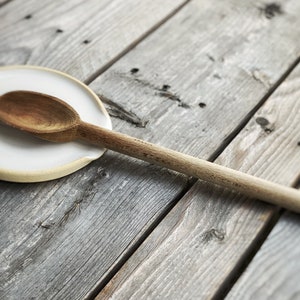 May include: A wooden spoon rests on a white ceramic spoon rest with a light brown rim. The spoon and rest are on a weathered, gray wooden surface, creating a rustic kitchenware scene.