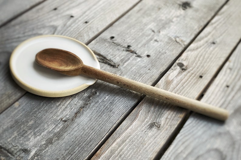 May include: A wooden spoon rests on a white ceramic spoon rest with a light brown rim. The spoon and rest are on a weathered, gray wooden surface, creating a rustic kitchenware scene.