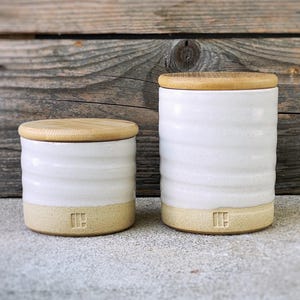 May include: Two white ceramic canisters with wooden lids. The canisters have a textured, wavy design and a light brown base. The smaller canister is on the left and the larger canister is on the right.