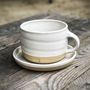 May include: White ceramic mug with a brown rim and a matching saucer. The mug has a slightly rounded shape and a handle on the right side.