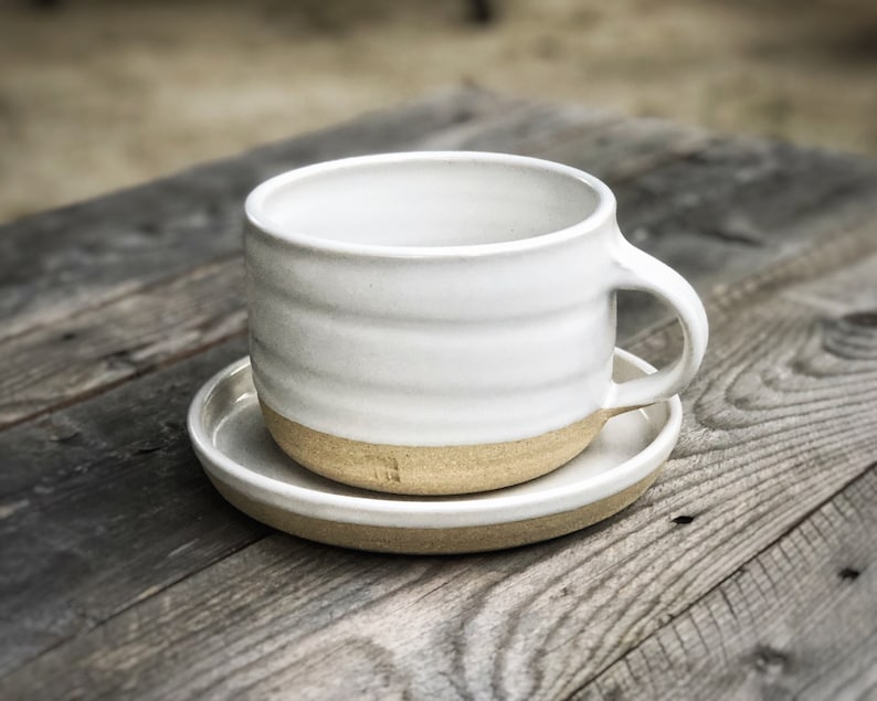 May include: White ceramic mug with a brown rim and a matching saucer. The mug has a slightly rounded shape and a handle on the right side.