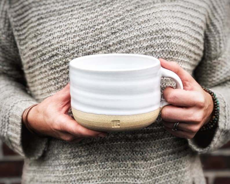May include: A white ceramic mug with a beige rim and a brown handle. The mug has a simple, elegant design and is perfect for enjoying a hot drink.