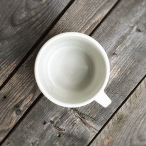 May include: A white ceramic mug with a rounded shape and a handle. The mug is empty and sitting on a wooden surface.