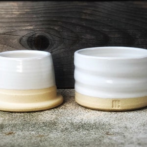 May include: Two white ceramic butter dishes with a light brown rim. The smaller dish has a smooth surface and the larger dish has a textured surface.