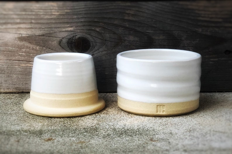 May include: Two white ceramic butter dishes with a light brown rim. The smaller dish has a smooth surface and the larger dish has a textured surface.