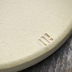 May include: Close-up of a round, off-white ceramic object with a textured surface. A small, rectangular stamp with three vertical lines and a horizontal line is visible on the edge. The background is a blurred wooden surface.