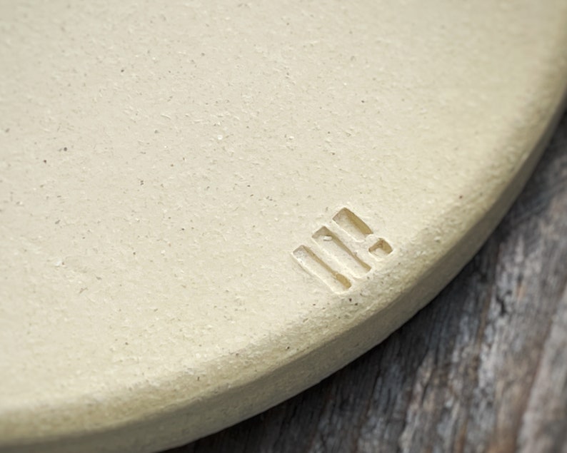 May include: Close-up of a round, off-white ceramic object with a textured surface. A small, rectangular stamp with three vertical lines and a horizontal line is visible on the edge. The background is a blurred wooden surface.