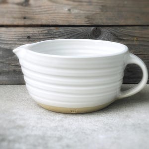 May include: White ceramic mixing bowl with a handle and a slightly raised rim. The bowl has a textured, ribbed design and a light brown base.