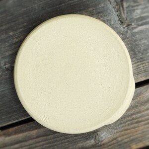 May include: A round, off-white ceramic spoon rest with a slightly textured surface. The spoon rest has a small indentation on one side and the initials "OR" are stamped on the bottom. The background is a weathered wooden surface.