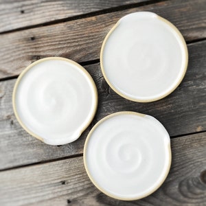 May include: Three white ceramic spoon rests with a natural, unglazed rim. The spoon rests have a circular shape with a small notch on one side. The background is a weathered wooden surface.