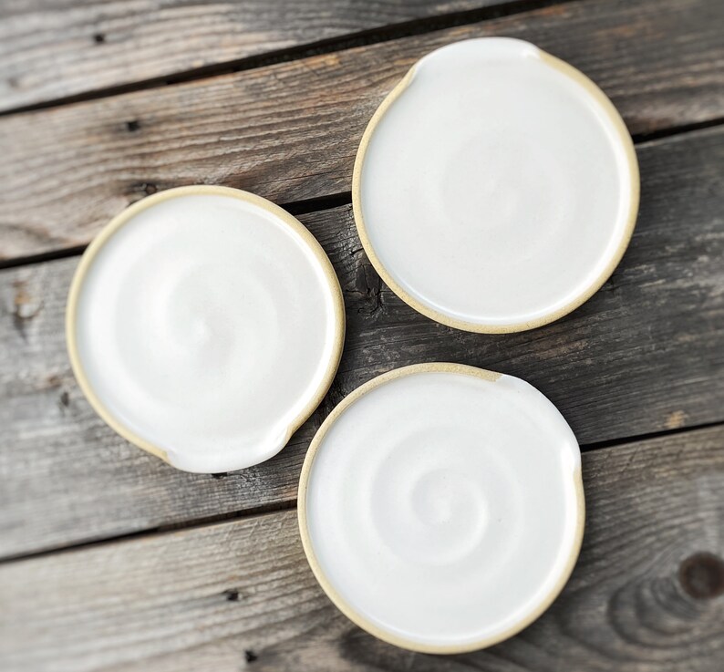 May include: Three white ceramic spoon rests with a natural, unglazed rim. The spoon rests have a circular shape with a small notch on one side. The background is a weathered wooden surface.
