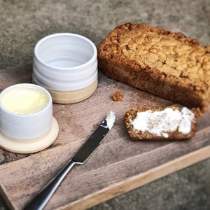May include: A wooden cutting board with a loaf of bread, a butter dish with butter, a butter knife, and a piece of bread with butter on it.