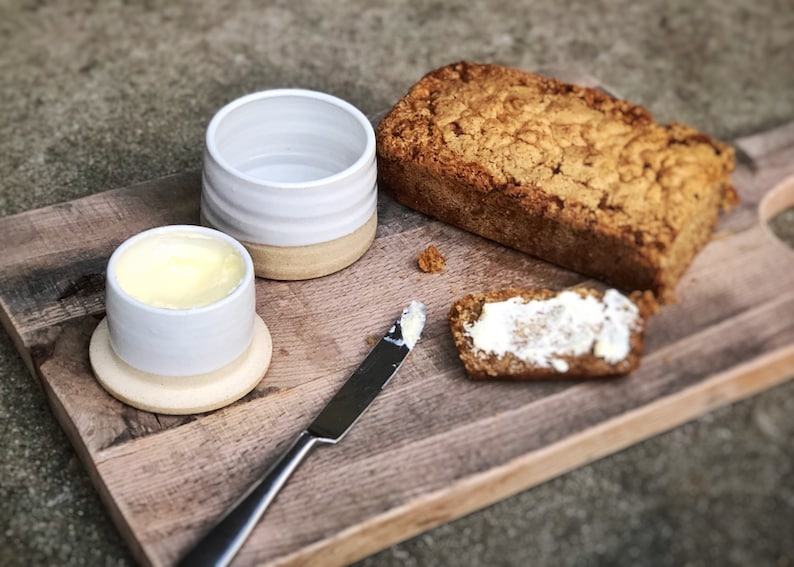 May include: A wooden cutting board with a loaf of bread, a butter dish with butter, a butter knife, and a piece of bread with butter on it.
