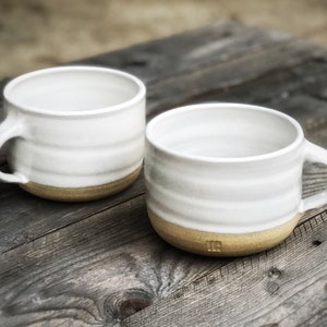May include: Two white ceramic mugs with a brown rim. The mugs have a slightly textured, wavy design.