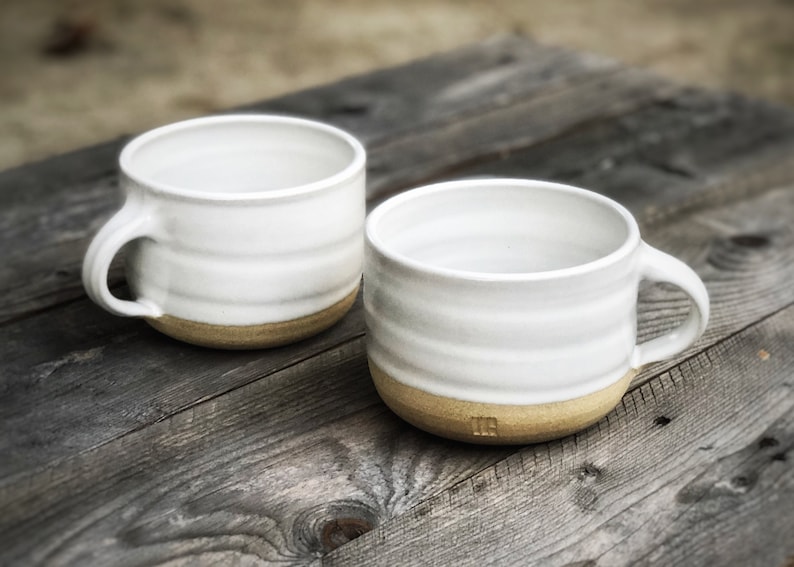 May include: Two white ceramic mugs with a brown rim. The mugs have a slightly textured, wavy design.