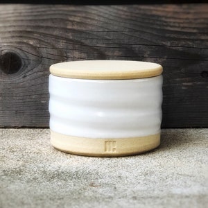 May include: White ceramic jar with a lid and a light brown rim. The jar has a textured, wavy design and the lid has a smooth surface. The jar has the letters "IH" on the bottom.