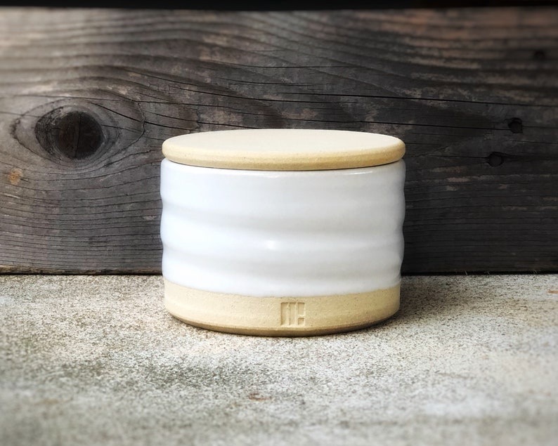 May include: White ceramic jar with a lid and a light brown rim. The jar has a textured, wavy design and the lid has a smooth surface. The jar has the letters "IH" on the bottom.