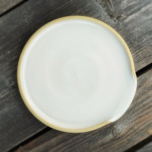 May include: A white ceramic spoon rest with a light tan rim. The spoon rest has a circular shape with a small indentation on one side. The spoon rest is on a weathered wooden surface.