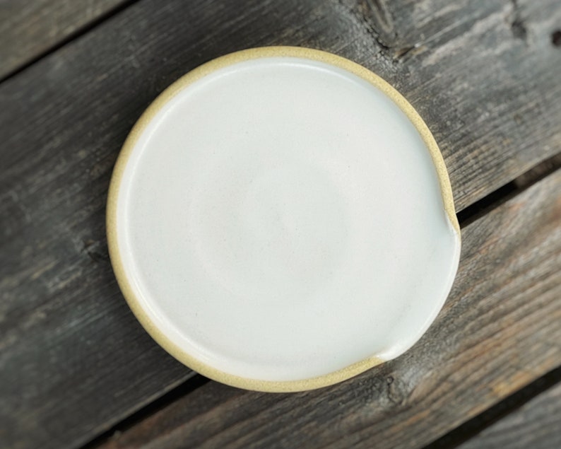 May include: A white ceramic spoon rest with a light tan rim. The spoon rest has a circular shape with a small indentation on one side. The spoon rest is on a weathered wooden surface.