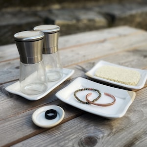 May include: Two silver salt and pepper shakers on a white rectangular dish, a white rectangular dish with a brown bracelet and a green beaded bracelet, and a small white dish with a black ring.