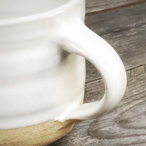 May include: White ceramic mug with a rounded handle and a light brown rim. The mug is sitting on a wooden surface.