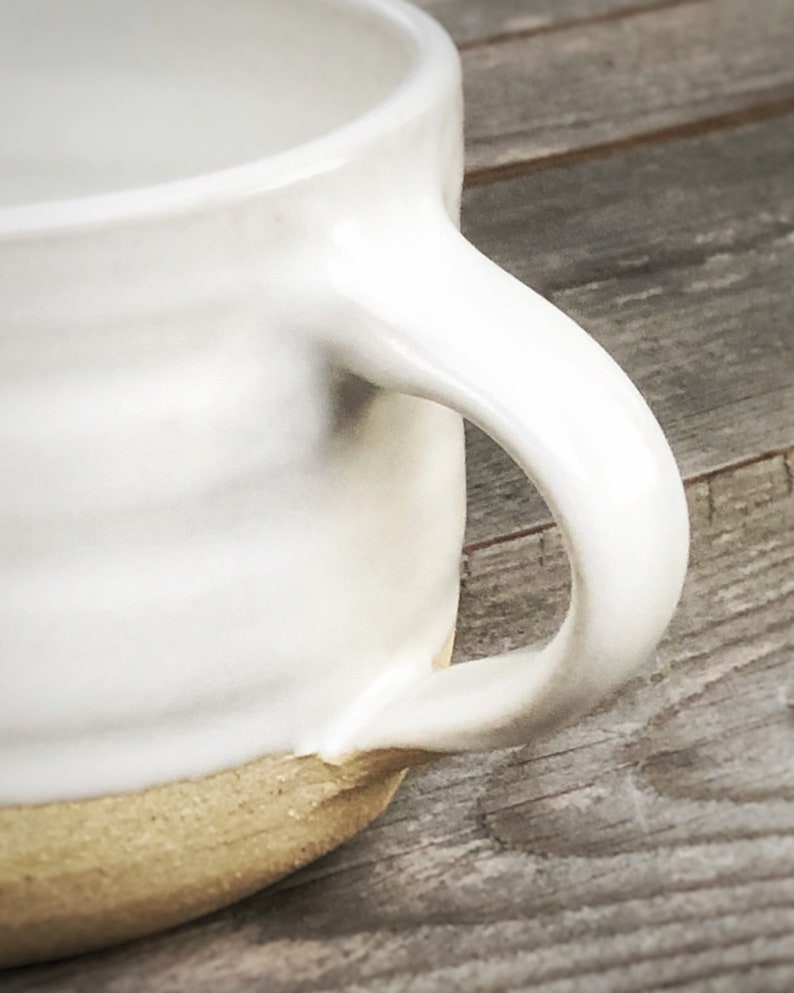 May include: White ceramic mug with a rounded handle and a light brown rim. The mug is sitting on a wooden surface.