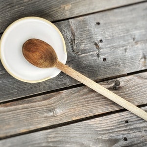 May include: A wooden spoon rests on a white ceramic spoon rest with a light brown rim. The spoon is a medium brown color and has a light-colored handle. The spoon rest is on a weathered wooden surface.