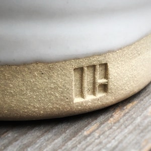 May include: Close-up of a white ceramic mug with a light brown rim. The mug has a textured bottom and the letters "IH" are stamped into the bottom.