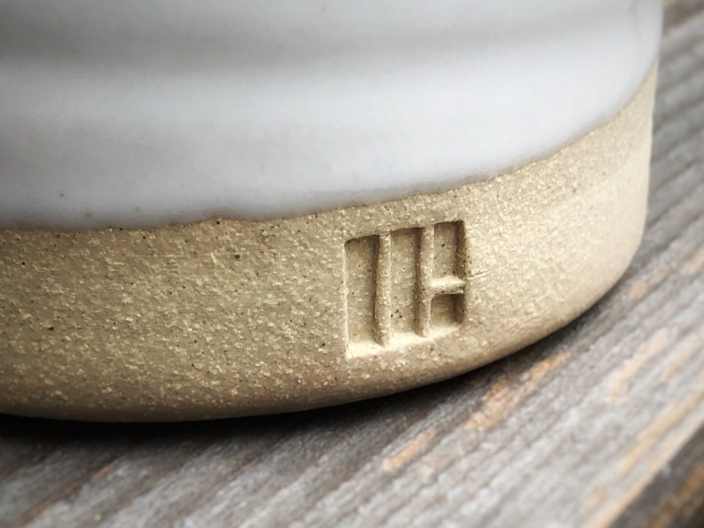 May include: Close-up of a white ceramic mug with a light brown rim. The mug has a textured bottom and the letters "IH" are stamped into the bottom.