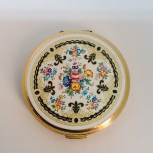 Op de afbeelding: Een ronde, goudkleurige compactspiegel met een witte achtergrond en een bloemenmotief. Het ontwerp heeft een centraal boeket van roze, gele en blauwe bloemen omringd door kleinere bloemenstekjes. Het bloemenmotief is omlijst door een zwarte en gouden rand.
