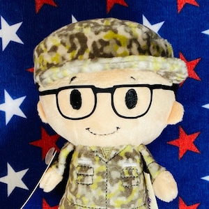 May include: A plush doll of a bespectacled character wearing a camouflage hat and jacket. The doll has a smiling face and is set against a blue background with red and white stars.