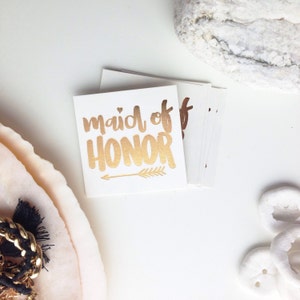 May include: Gold foil temporary tattoo with the text "maid of honor" and a small arrow. The tattoo is on a white background.