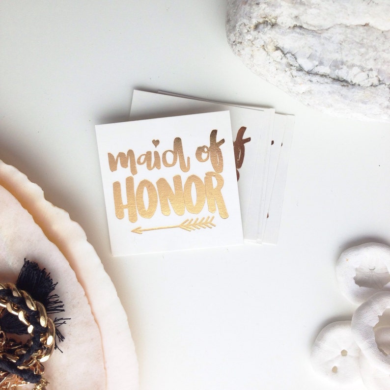 May include: Gold foil temporary tattoo with the text "maid of honor" and a small arrow. The tattoo is on a white background.